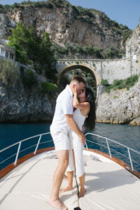 Newly engaged couple hugging and smiling on a yacht beneath the iconic Amalfi cliff bridge at golden hour