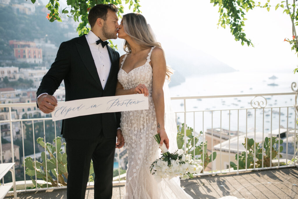 Bride and groom kissing on a Positano terrace overlooking the Amalfi Coast, holding a sign that reads “Eloped in Positano” — romantic Italian elopement with sea views and Mediterranean charm